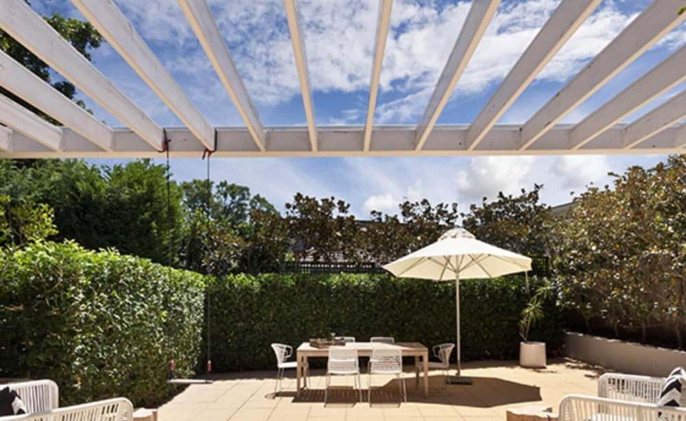 Blik vanaf een terras onder een witte houten pergola met heldere platen, met daarachter een tuin met eettafel en parasol