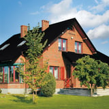 Bakstenen huis met dak van dakpanplaat Szafir 350/15 Gitzwart, 0,50 mm, omringd door bomen