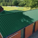 Dak van damwandplaat 35/207 Mosgroen op een bakstenen gebouw naast een sportveld
