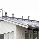 Zwart Weckman looprooster gemonteerd op een dak met dakpannen, naast schoorstenen en ventilatieopeningen