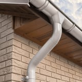 Detail van een zilver-metallic regenpijp met bocht en beugel, gemonteerd aan een bakstenen muur