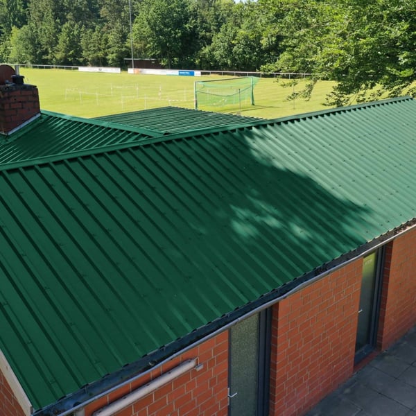 Dak van damwandplaat 35/207 Mosgroen op een bakstenen gebouw naast een sportveld
