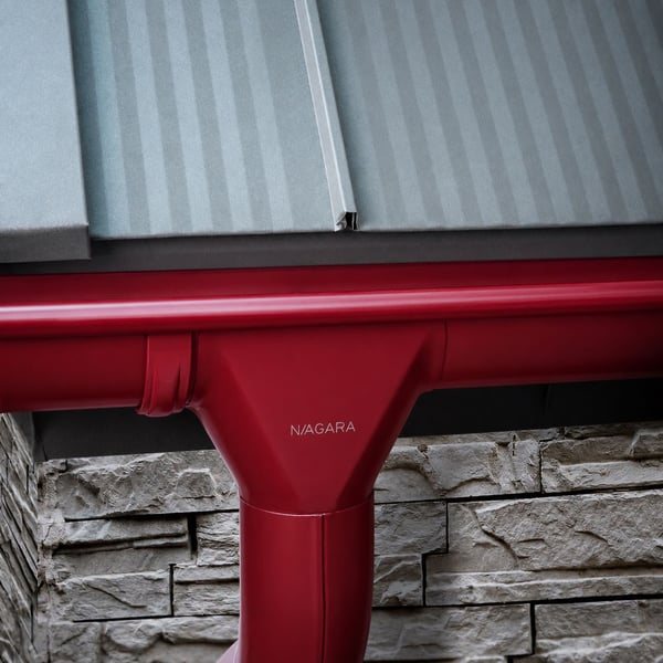 Detail van kersrode (RAL 3011) dakgootuitloop met Niagara logo, gemonteerd onder een lichtgrijs dak