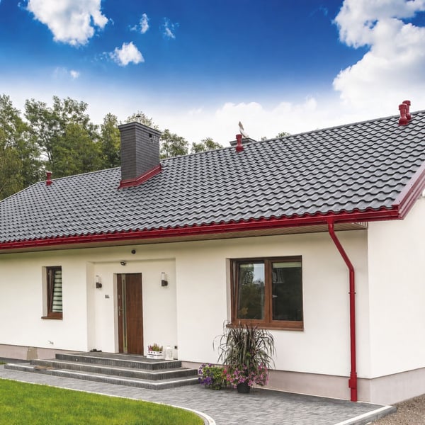 Kersrode regenpijpbeugel gemonteerd aan een wit huis met donker dak en rode dakgoten