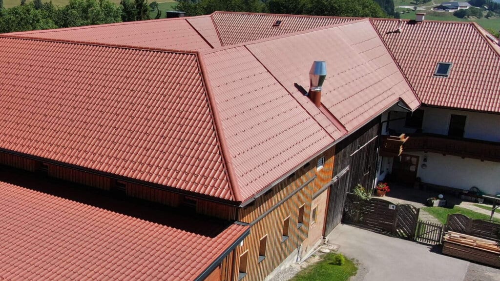 Landelijk gebouwencomplex met grote daken van rood gecoate dakpanplaat in baksteenlook, gecombineerd met houten gevels, gelegen in een heuvelachtig landschap met uitzicht op groene weiden en bossen.