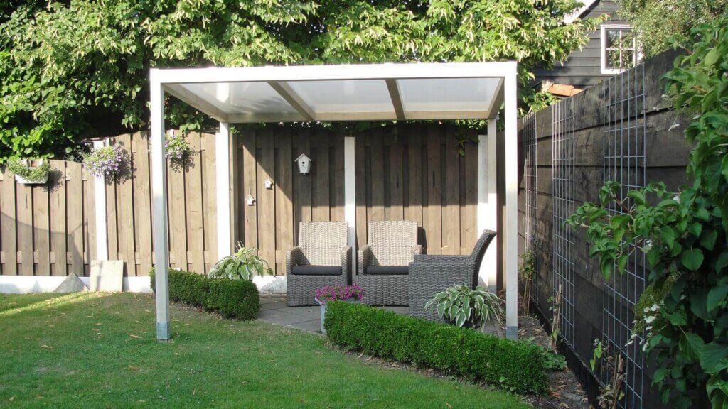 Terrasoverkapping van aluminium met heldere kunststof dakbedekking in een verzorgde tuin, met daaronder een zitgroep van polyrotan, omgeven door een houten schutting en beplanting.