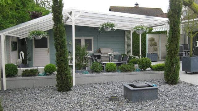 Ruime terrasoverkapping van wit aluminium met heldere dakbedekking, geïntegreerd in een modern aangelegde tuin met siergrind, verhoogde plantenbakken, vormsnoei en tuinmeubelen voor een lichtblauw tuinhuisje.