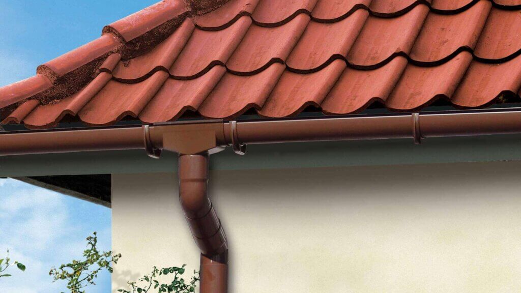 Close-up van een huisdak met klassieke rode dakpannen en bruine halfronde kunststof dakgoot, inclusief regenpijp en verbindingsstuk aan de druiplijst.